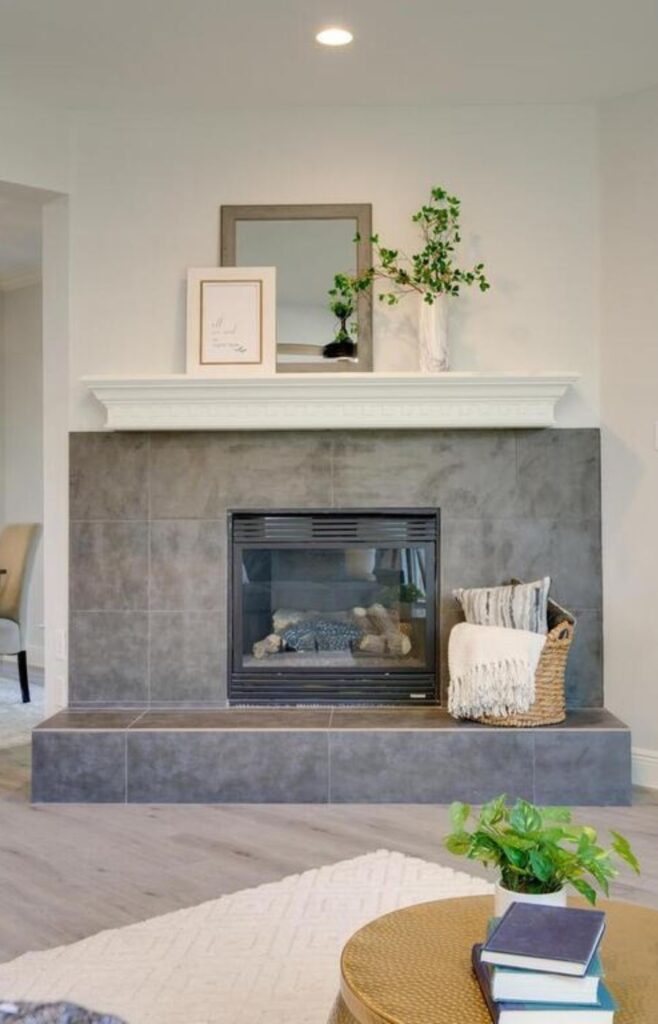 A stylish and cozy grey tiled fireplace with a white mantle highlighted by a mirror, a picture, a blanket basket, and a vibrant green plant. The grey tiles add a modern touch, while the white mantle provides a clean contrast. The mirror and picture enhance the visual interest of the space, while the blanket basket offers a practical and decorative element. The green plant brings a pop of color and life to the setting, creating a welcoming and inviting ambiance in the room.