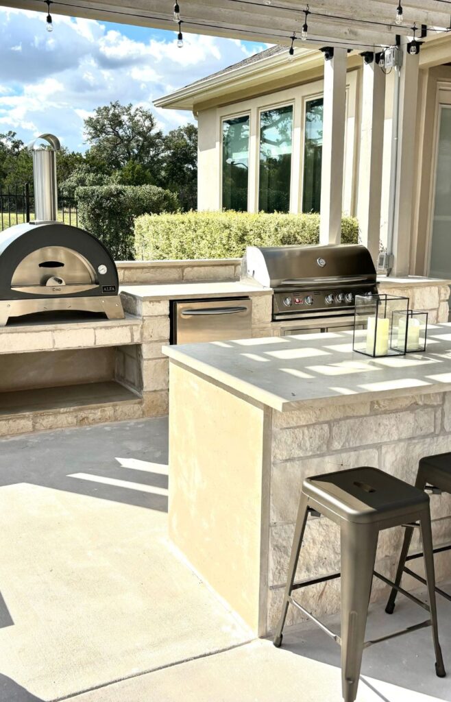 A charming outdoor kitchen designed with natural stone elements, featuring a built-in barbecue grill and a traditional pizza fire oven as its focal points. The rustic appeal of the stone construction blends beautifully with the surrounding landscape, creating a cozy and inviting outdoor cooking area.
