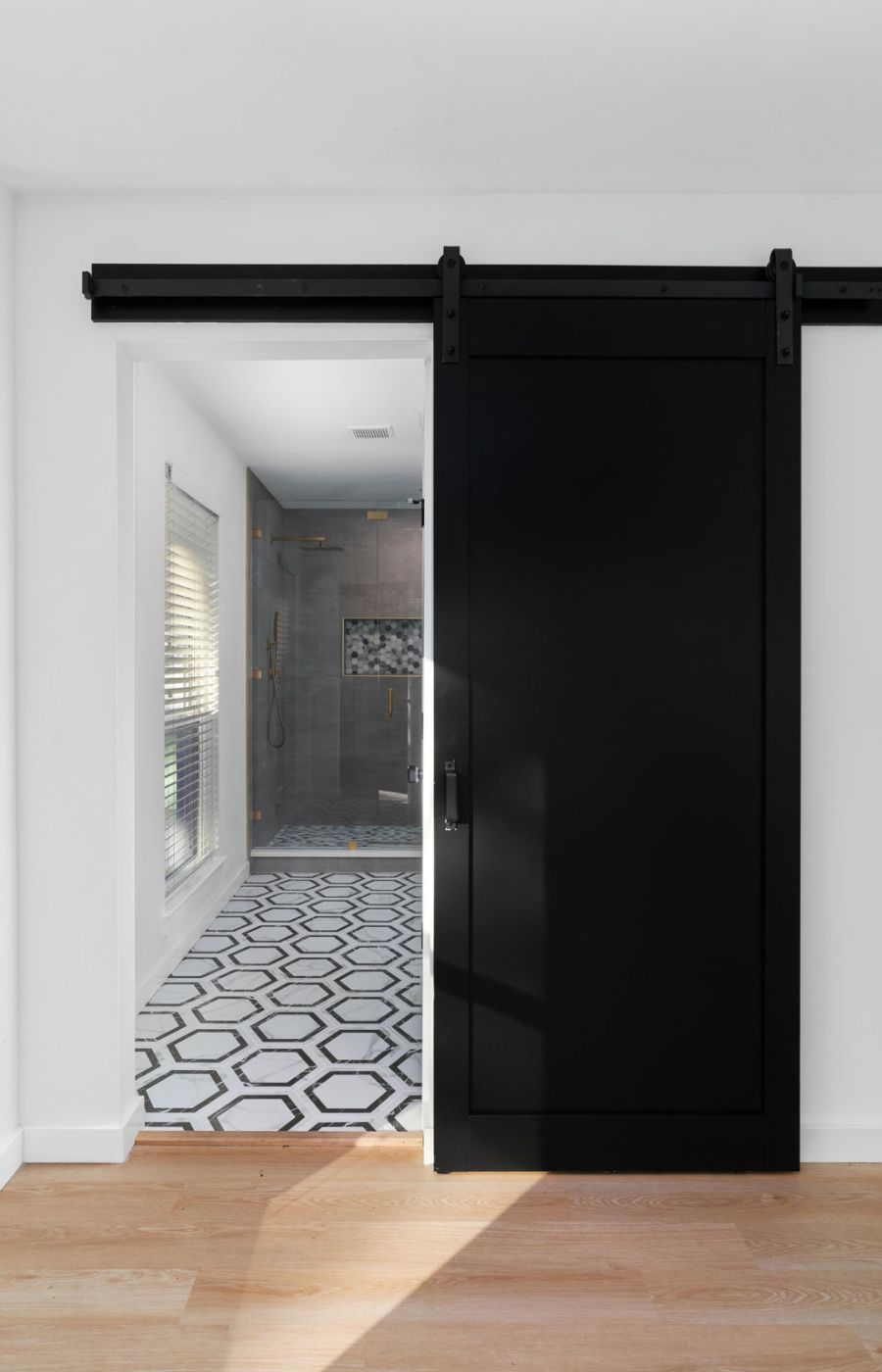 A striking black barn door in the bathroom, adds a bold and rustic touch to the space. The door stands out against the neutral tones of the bathroom, creating a visually appealing contrast. Its design brings a sense of farmhouse charm and character to the modern setting, enhancing the overall aesthetic with a touch of industrial style. The black barn door serves as a unique and eye-catching feature, offering both functionality and visual interest in the primary room.